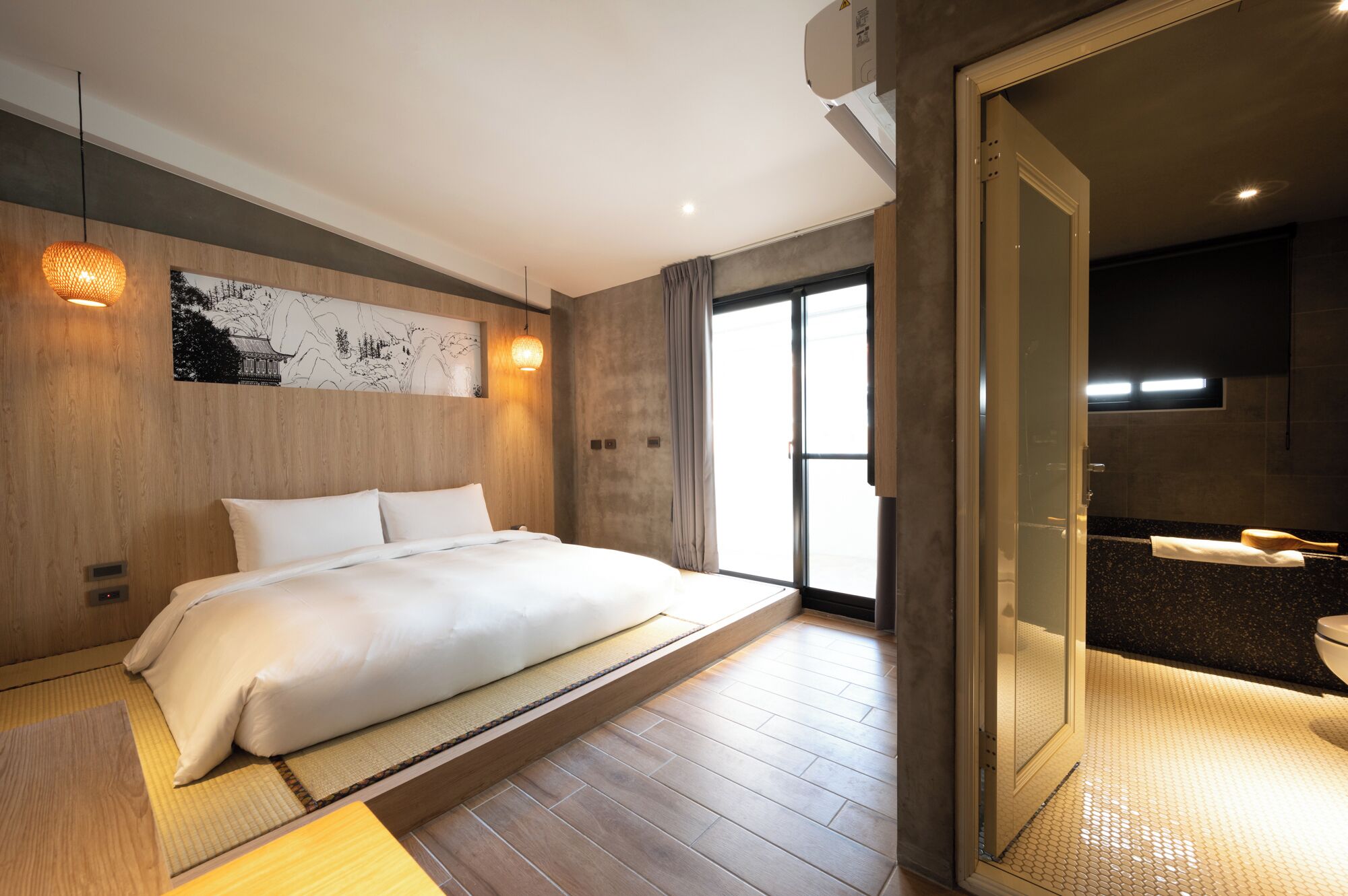 A modern hotel room with a large bed, a wooden accent wall, a large window, and a separate bathroom area.