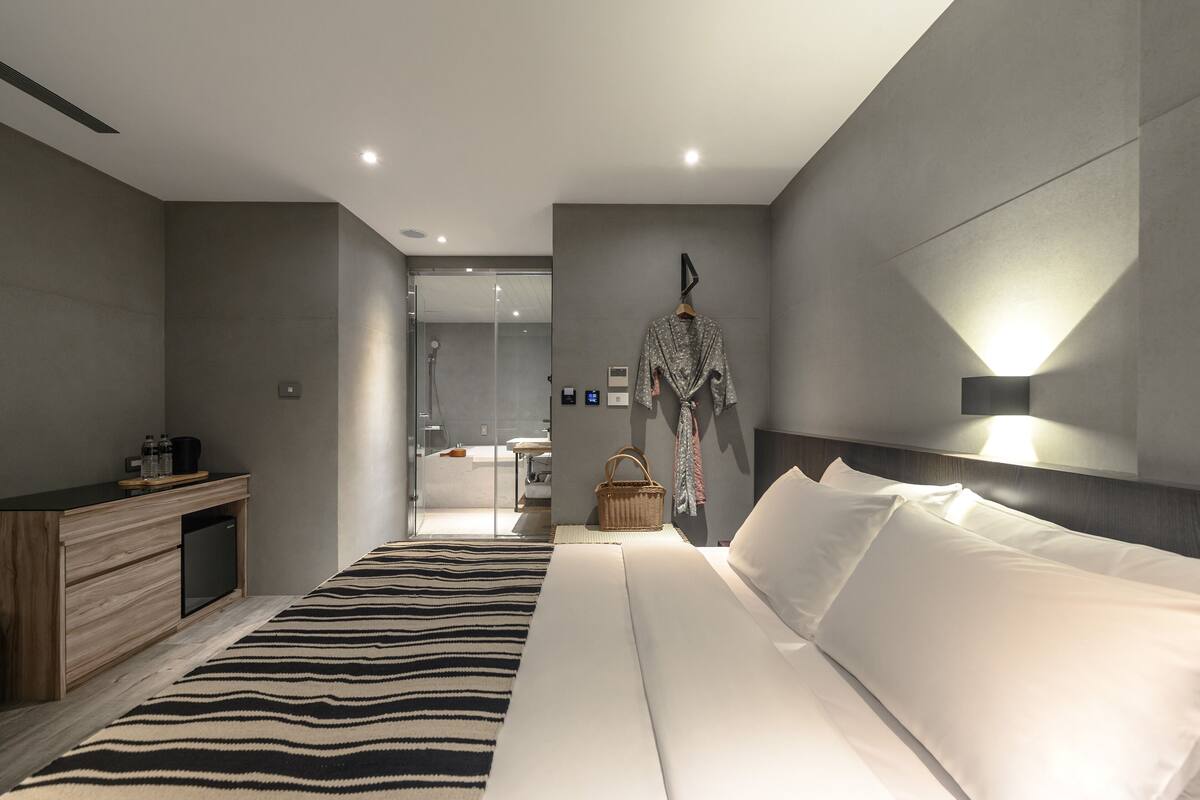 A modern hotel room with a large bed, a bedside table with a lamp, a small desk, and a shower area visible through a glass door.