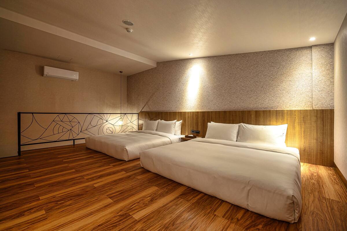 A hotel room with two beds, wooden flooring, a textured wall, and a ceiling light.