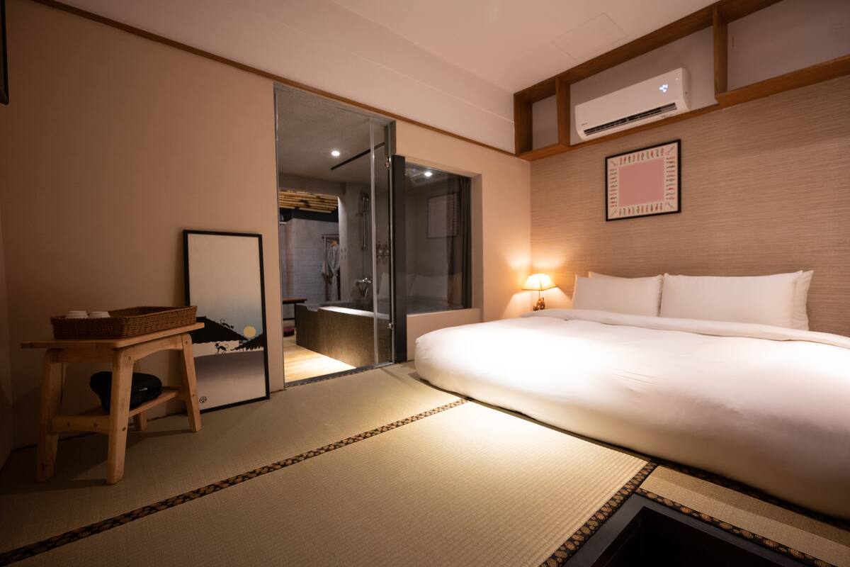 A hotel room with a bed, a mirror, a small table with a basket, a wall-mounted air conditioner, and a framed picture.
