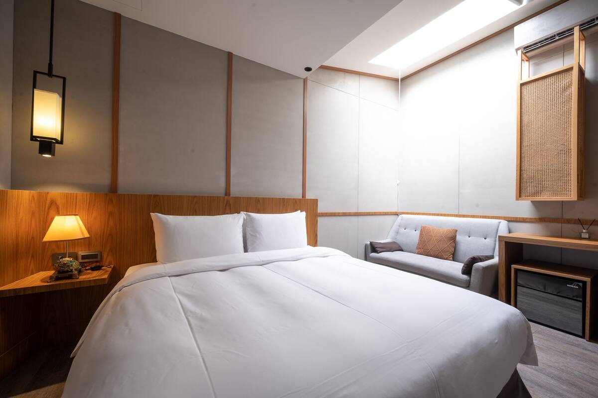 A hotel room with a large bed, a bedside table with a lamp, a small desk, and a sofa.