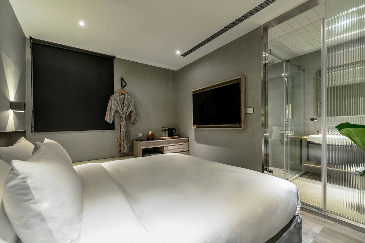 A modern hotel room with a large bed, a bedside table, a robe hanging on a hook, a mounted TV, and a glass-enclosed shower area.