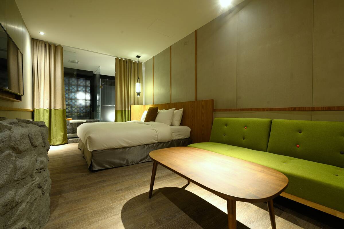 A hotel room with a bed, a green sofa, a wooden table, and a large window with curtains.