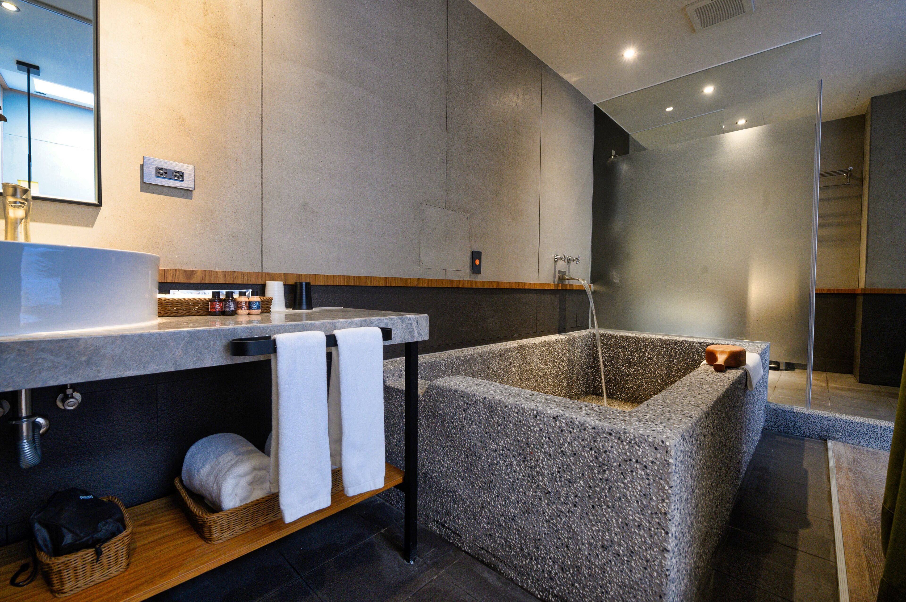 A modern bathroom with a large stone bathtub, a wooden shelf with towels, and a mirror above the sink.