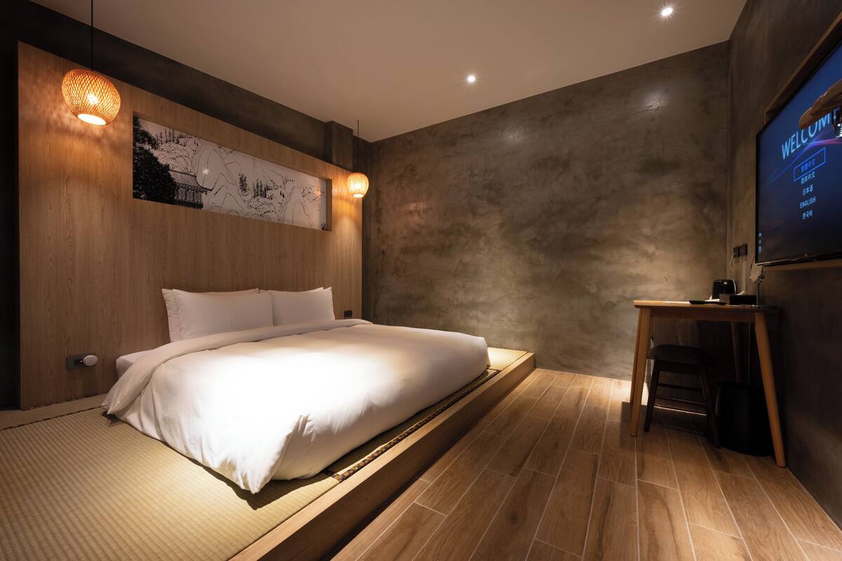A modern bedroom with a large bed, a wooden headboard, a wall-mounted TV, and a small table with a chair.