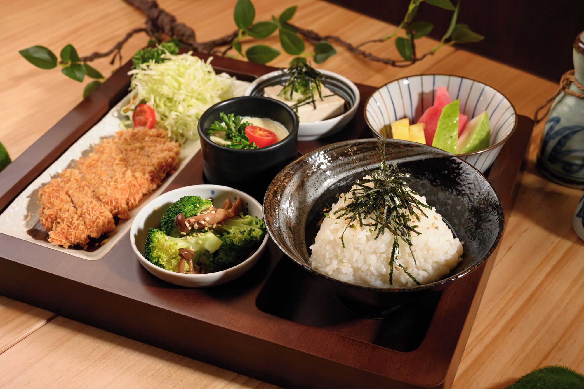 A wooden tray with various dishes including rice, broccoli, and a piece of breaded meat.