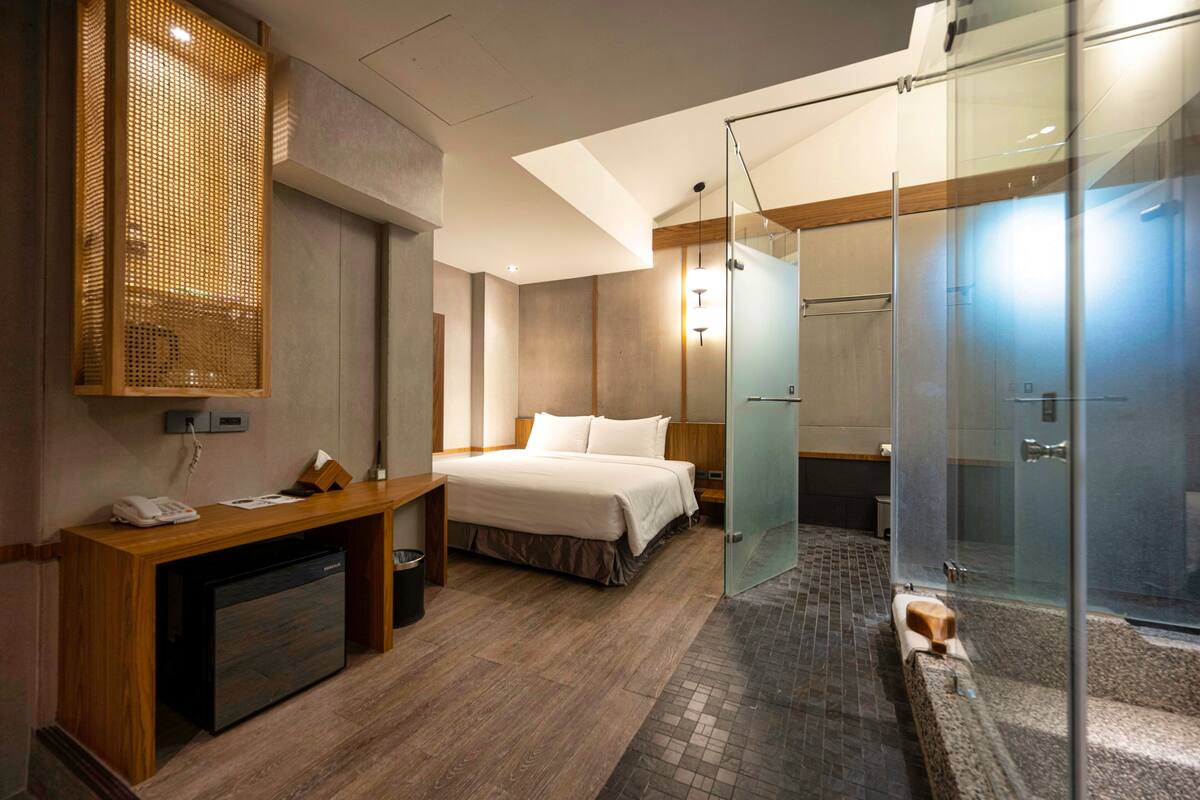 A modern hotel room with a bed, desk, and a glass-enclosed shower area.