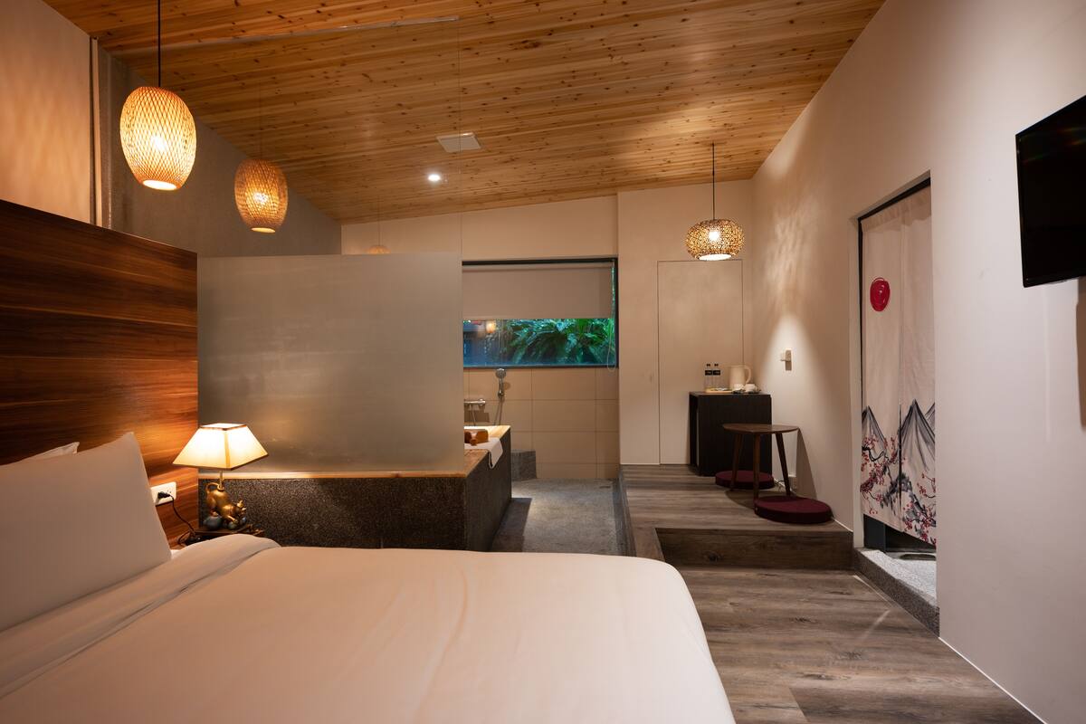 A modern hotel room with a wooden ceiling, a large bed, a desk, and a bathroom visible through an open door.