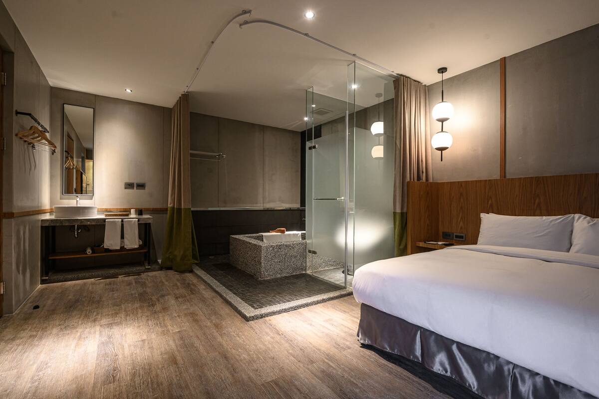 A modern hotel room with a large bed, a glass-enclosed shower, a vanity with a mirror, and a wooden headboard.