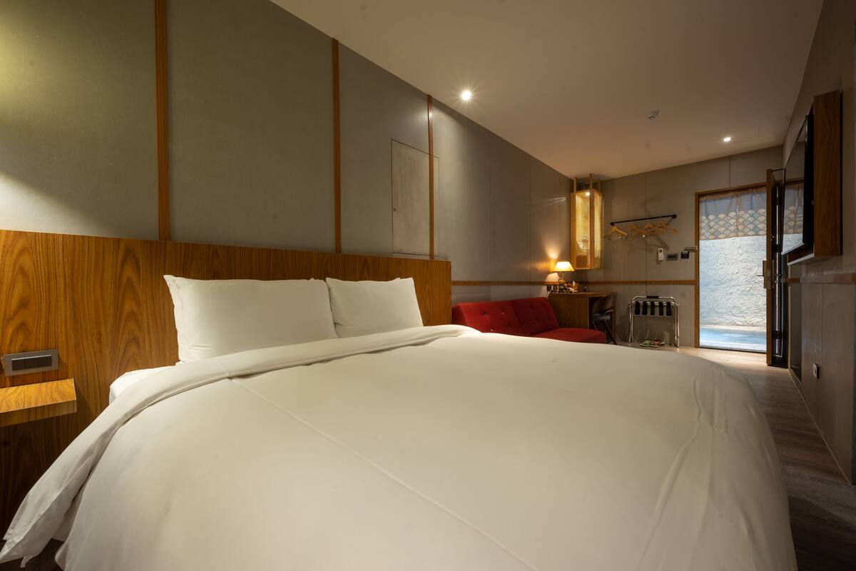 A hotel room with a large bed, a wooden headboard, a red chair, a small table, and a window with a view.