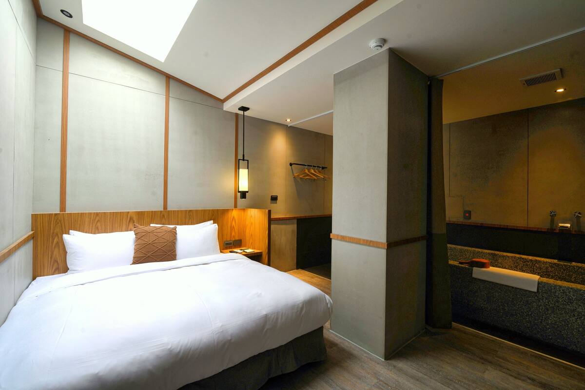 A hotel room with a large bed, a wooden headboard, a nightstand, a wall-mounted mirror, and a bathroom with a sink and towel rack.