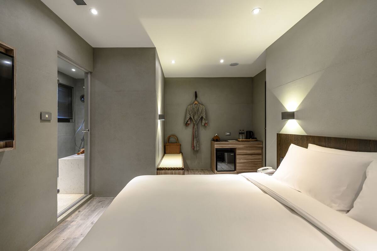 A modern hotel room with a large bed, a bedside table with a lamp, a small desk, and a wardrobe.