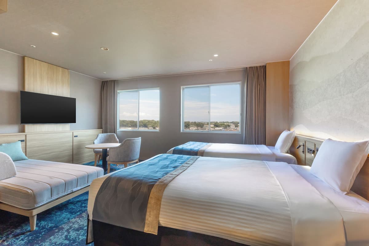 A hotel room with two beds, a flat-screen TV, a small table, and a view of the city.