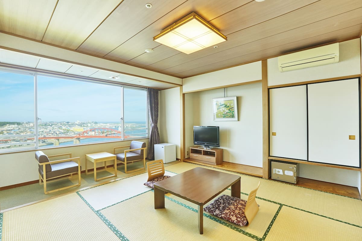 A hotel room with tatami flooring, a television, a small refrigerator, and a large window offering a city view.