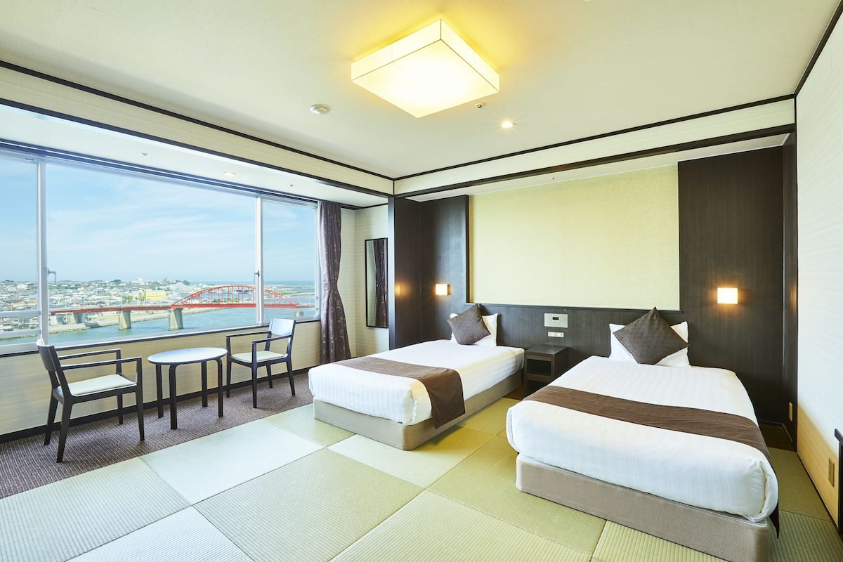A hotel room with two beds, a sitting area with chairs, a large window with a city view, and a modern design.