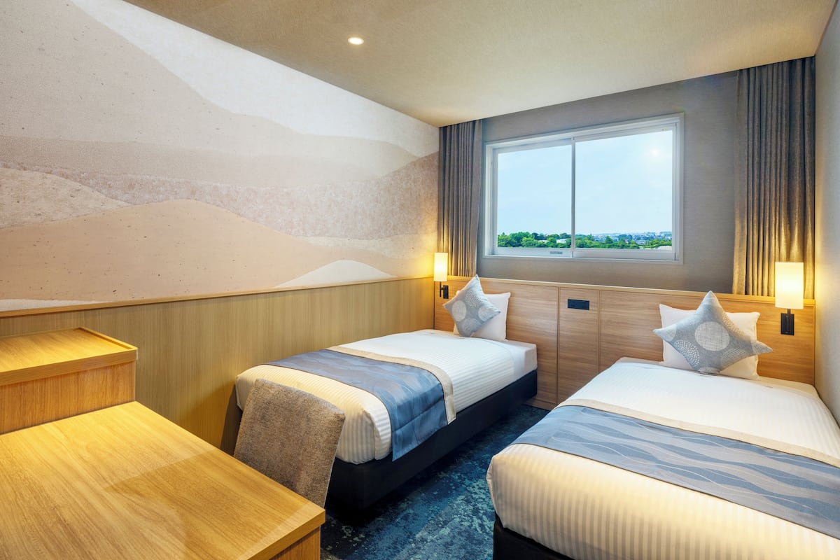 A hotel room with two beds, a desk, and a large window offering a city view.