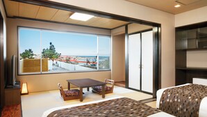 Habitación de hotel con un ventanal grande que ofrece vistas a la costa, una pequeña mesa con sillas y dos camas con ropa de cama estampada.