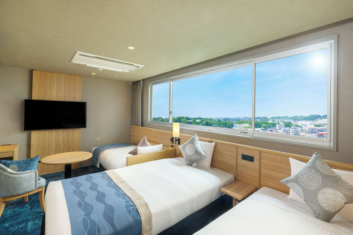 A hotel room with two beds, a TV, a window with a city view, and a small table.