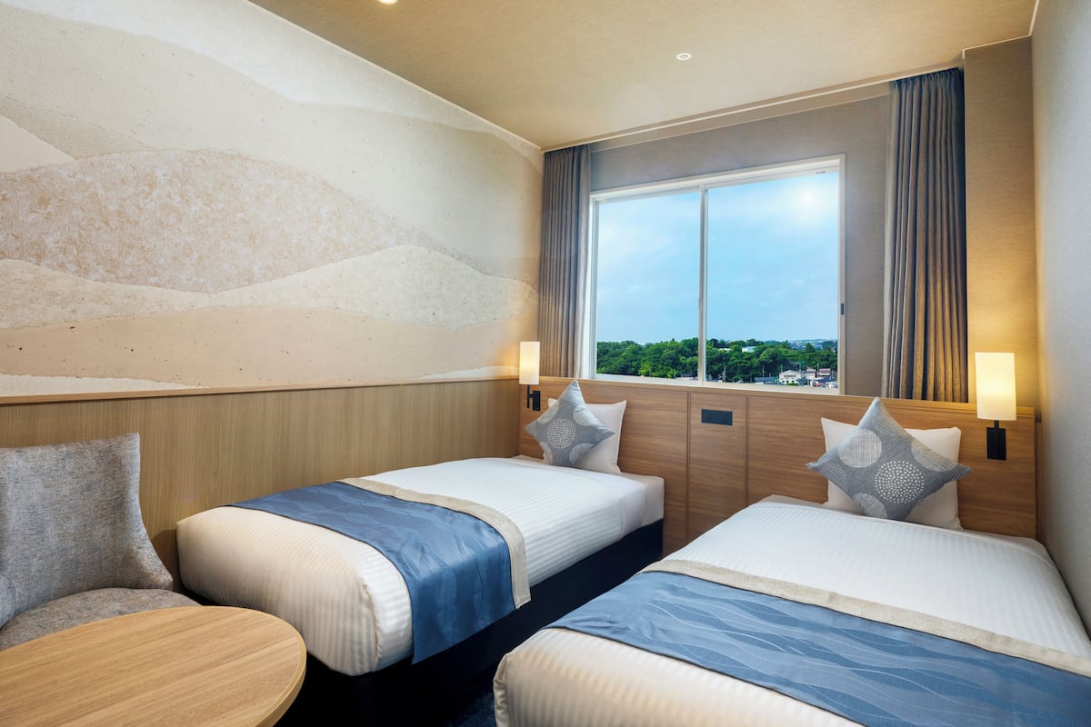 A hotel room with two beds, a large window with curtains, and a view of greenery outside.