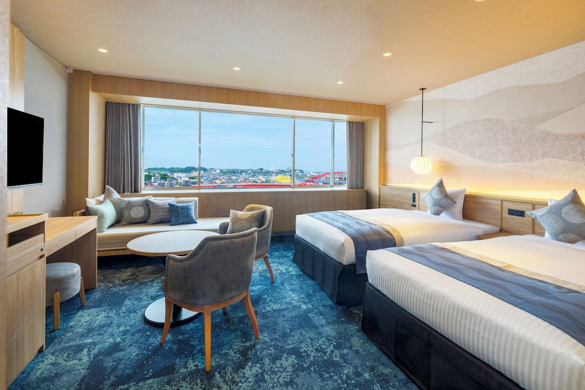A hotel room with a large window, a desk, two beds, and a seating area.