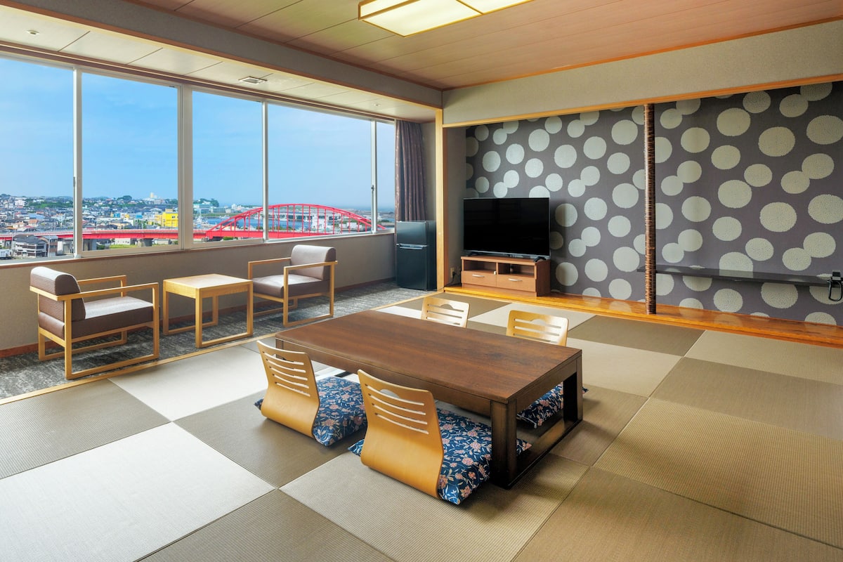 A room with tatami flooring, a wooden table, chairs, a television, and a large window with a view of a red bridge.