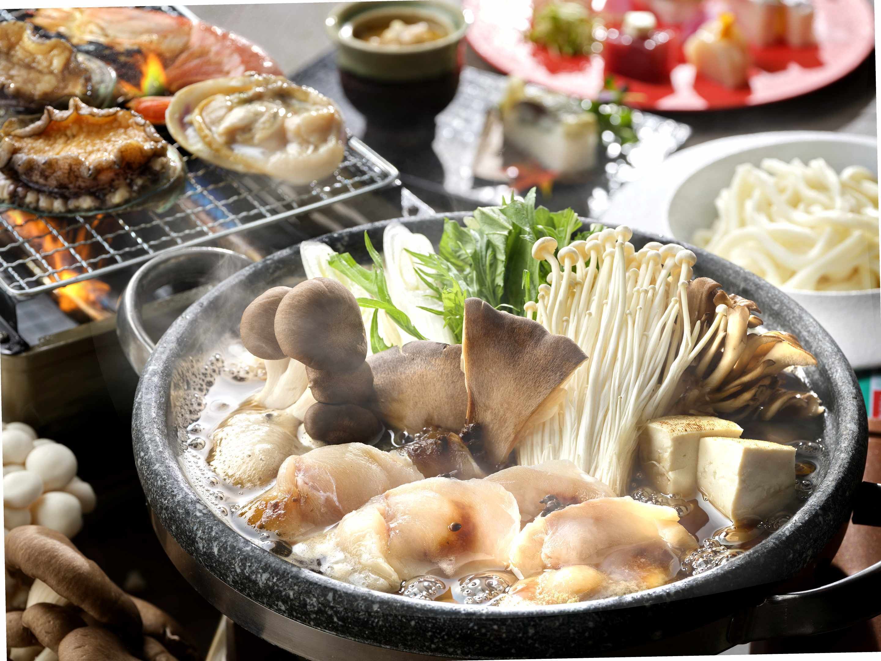 A hot pot with various ingredients like mushrooms and tofu, surrounded by other dishes and a grill.