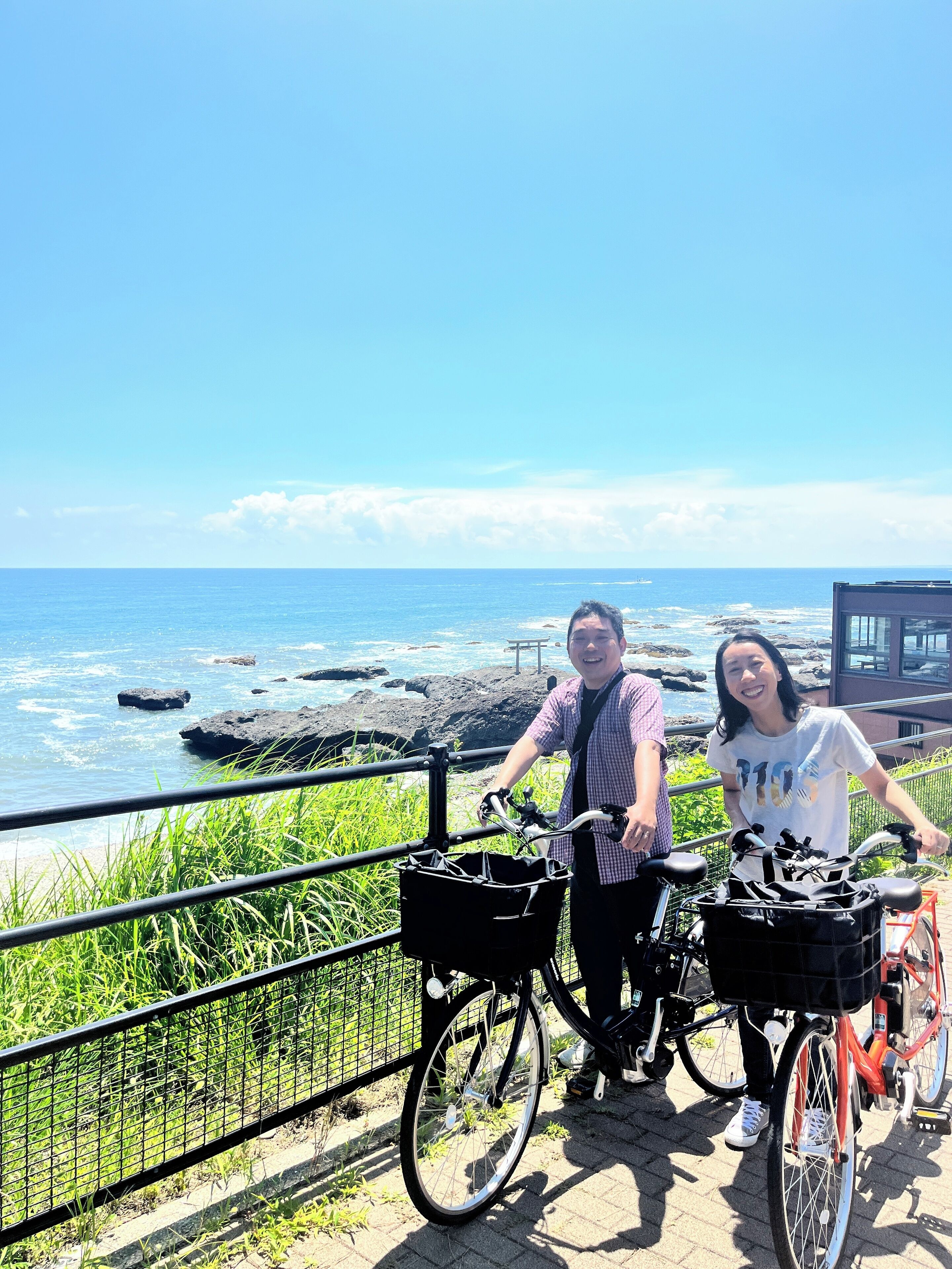 Two people standing by a railing with bicycles, overlooking a rocky coastline and the ocean.