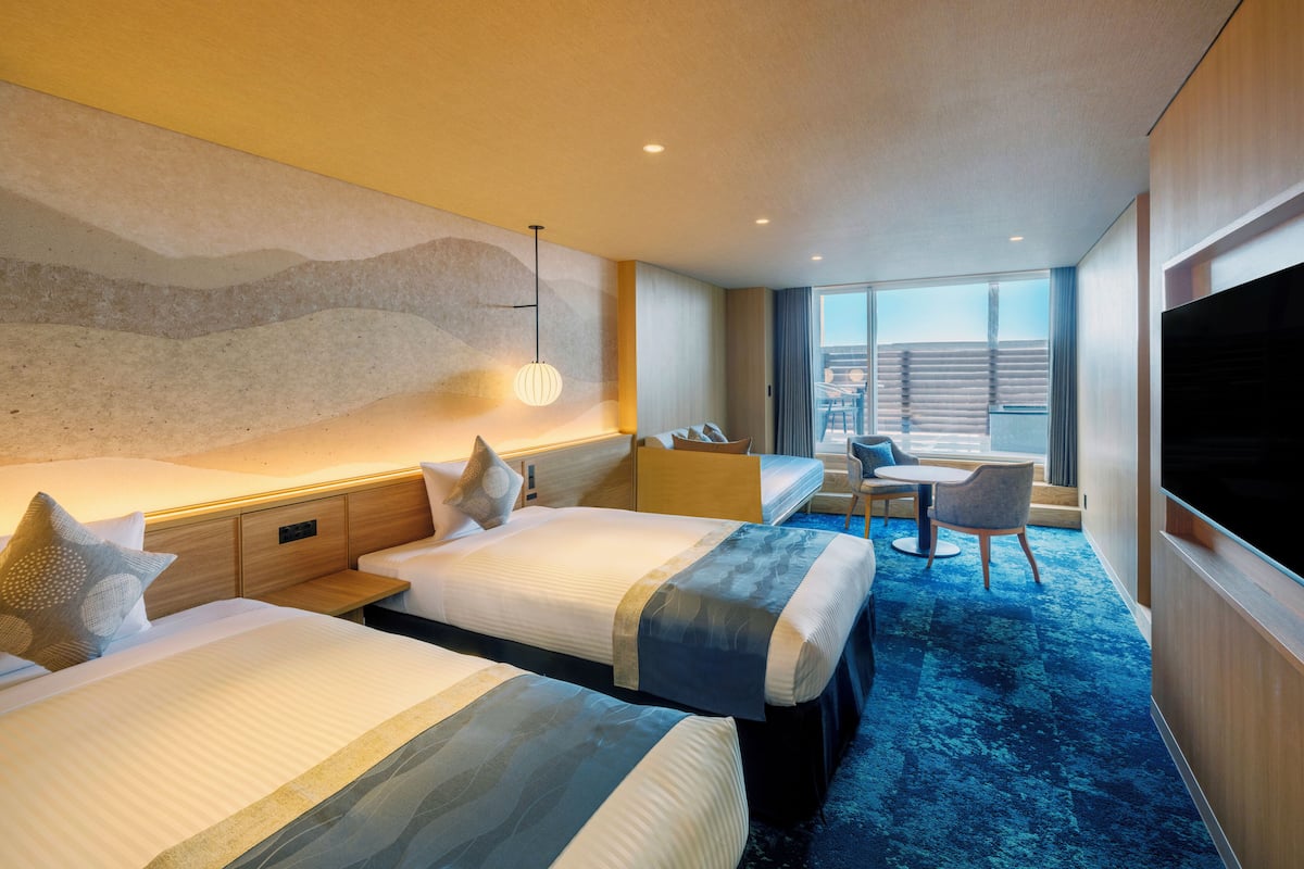 A hotel room with three beds, a TV, a desk with a chair, and a large window offering a view of the outside.