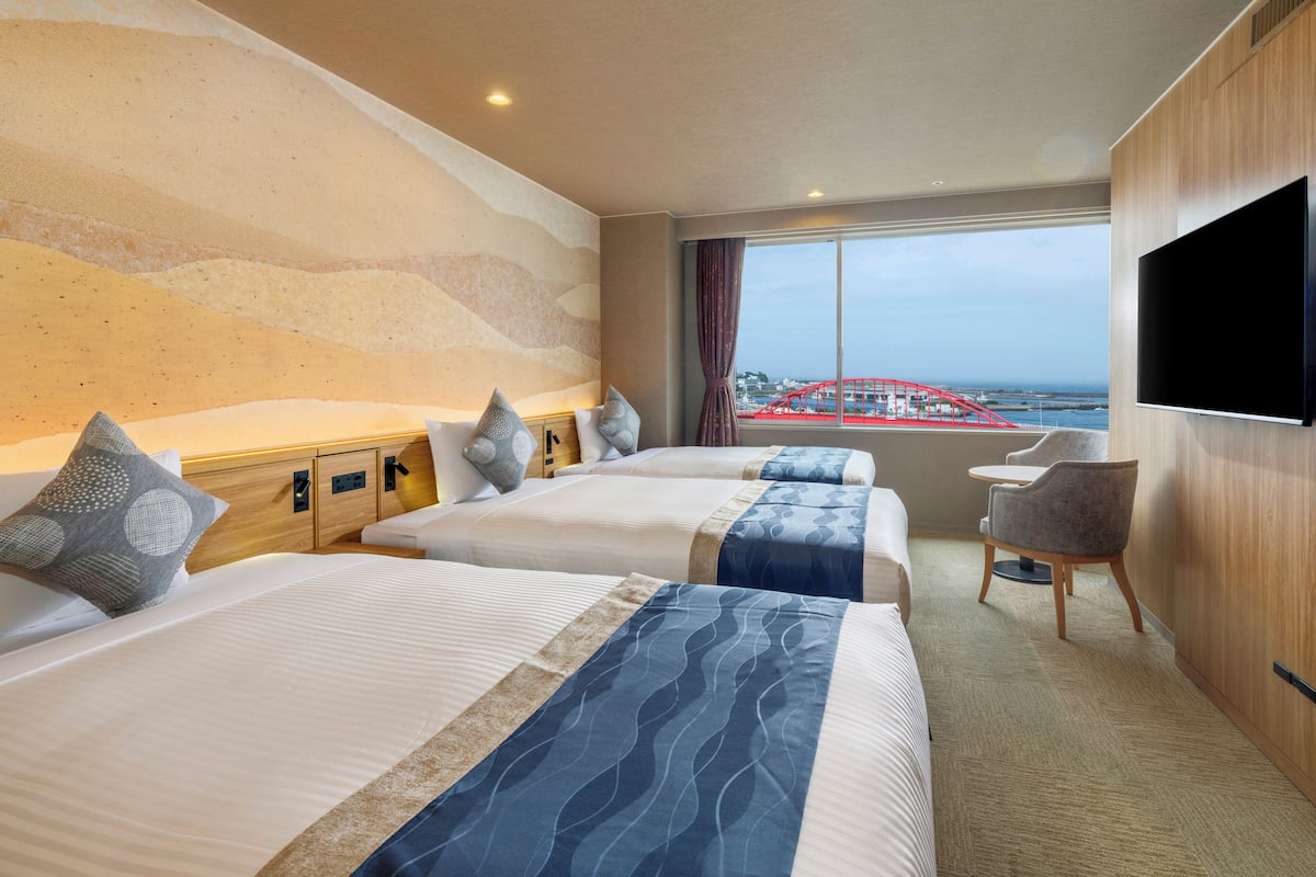 A hotel room with two beds, a TV, a desk, and a view of a red bridge.