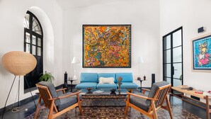 A modern living room with a blue sofa, wooden armchairs, a patterned rug, and a large abstract painting on the wall. - Casa Venus - Refine living in Historic District Mazatlan (Mazatlan)