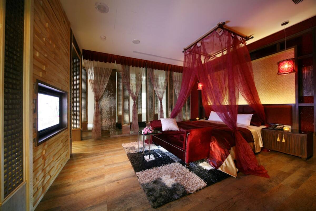 A modern bedroom with a wooden floor, a red canopy bed, a TV, and a nightstand.