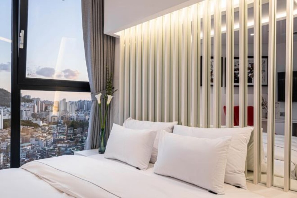 Quarto de hotel moderno com uma janela grande, cama com roupa de cama branca, painel decorativo na parede e vista para a cidade.