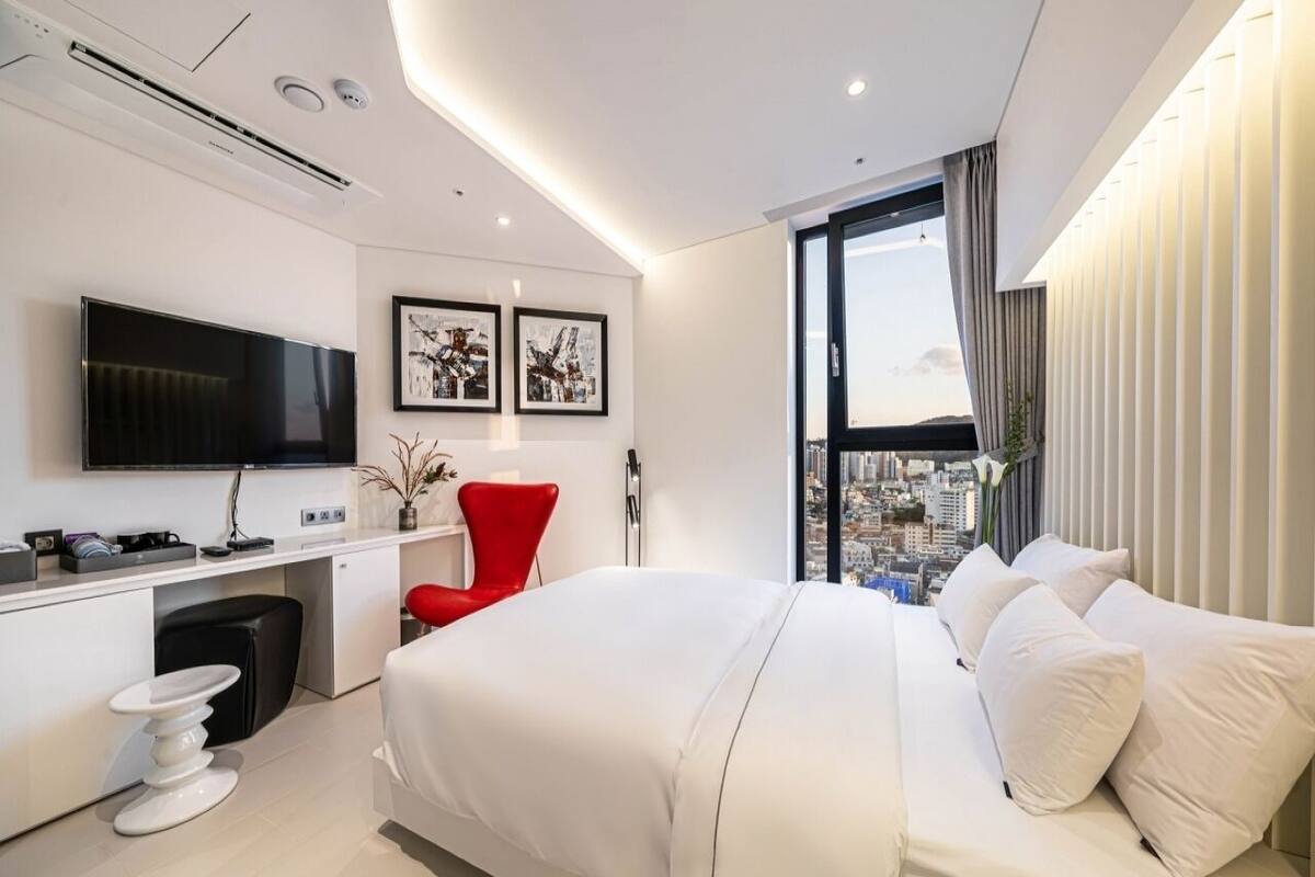 A modern hotel room with a large bed, a desk with a flat-screen TV, a red chair, and a city view through the window.