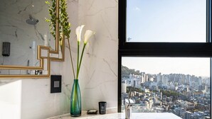 A modern bathroom with a large window, a marble countertop, a sink, and a vase with white flowers. - Forestar (Seoul)