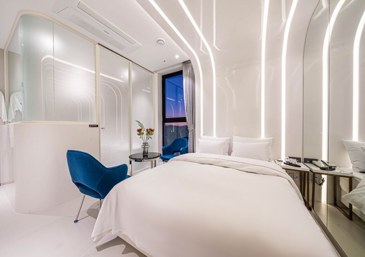 A modern hotel room with a large bed, a blue chair, a small table with flowers, and a view of the city at night.