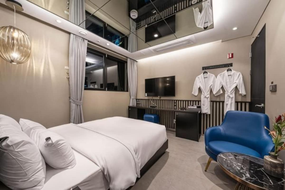 A modern hotel room with a bed, a blue armchair, a flat-screen TV, and a mirror.