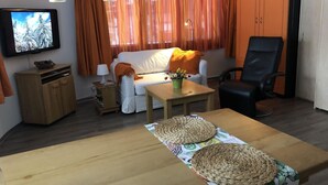 Ein Wohnzimmer mit einer Couch, einem Fernseher, einem Holztisch mit gewobenen Untersetzern, einem schwarzen Bürostuhl und orangefarbenen Vorhängen.