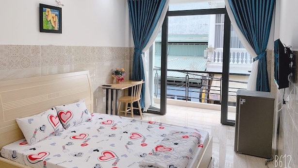 A hotel room with a bed, a small table with a vase of flowers, a desk, and a balcony with a view. - Song Huong House (Ho Chi Minh City)