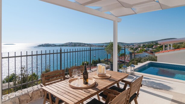 Une table en bois entourée de chaises, une bouteille et des verres, avec une vue dégagée sur une mer bleue et une piscine.