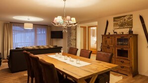 A dining area with a wooden table, brown chairs, and a chandelier. There is a sofa and a cabinet with glass doors in the background.