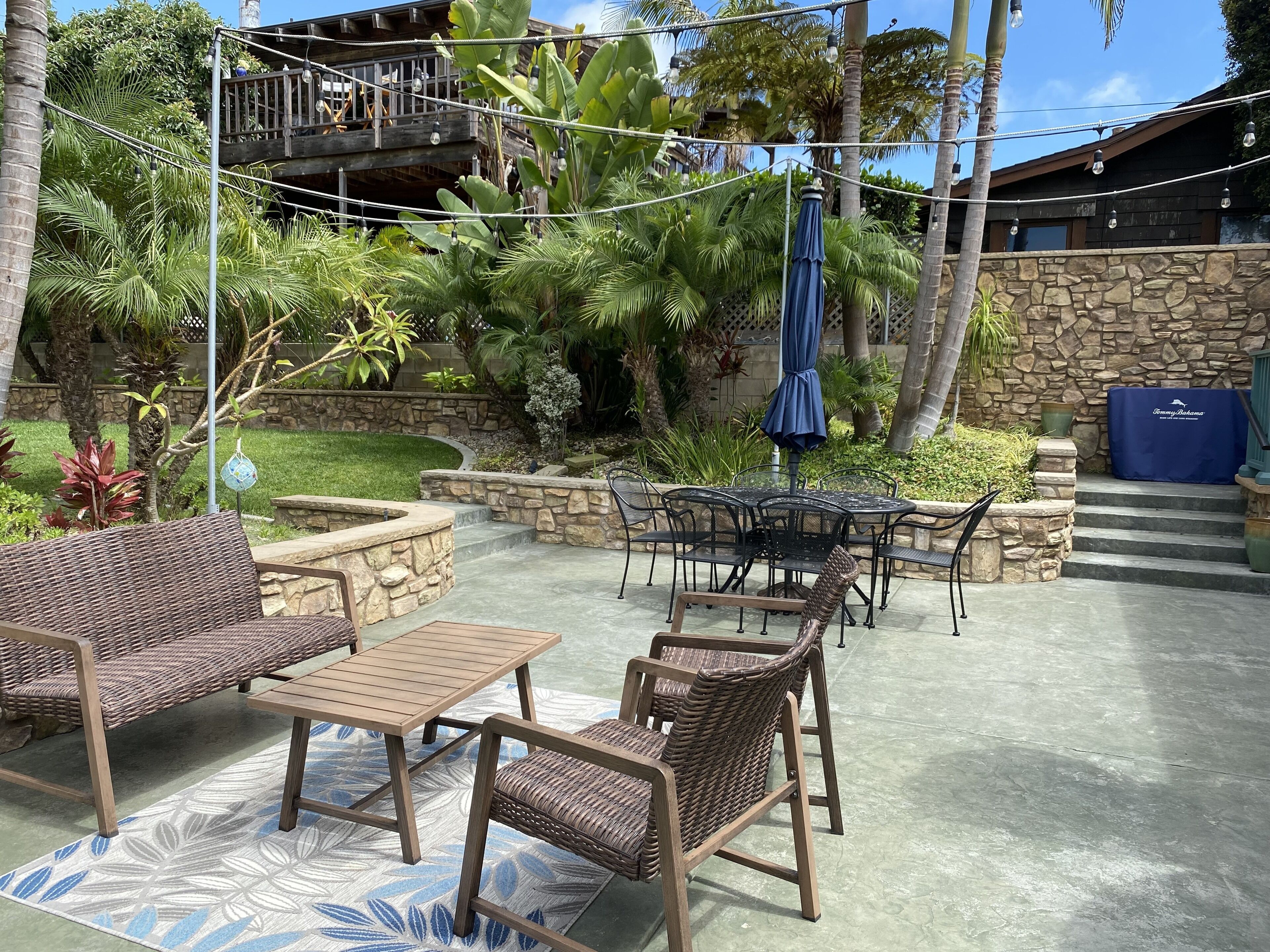 An outdoor patio area with wicker furniture, a stone fire pit, and a view of a wooden deck and lush greenery.