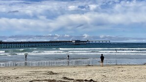 Ein Strand mit einem Steg, der sich ins Meer erstreckt, Menschen schwimmen und spazieren am Ufer entlang.