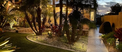 Un jardín bien cuidado con un sendero de piedra, palmeras y luces colgantes.