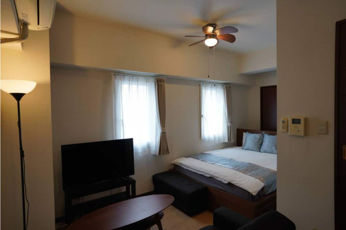 A hotel room with a bed, a television, a desk, a chair, a ceiling fan, and a window with curtains.