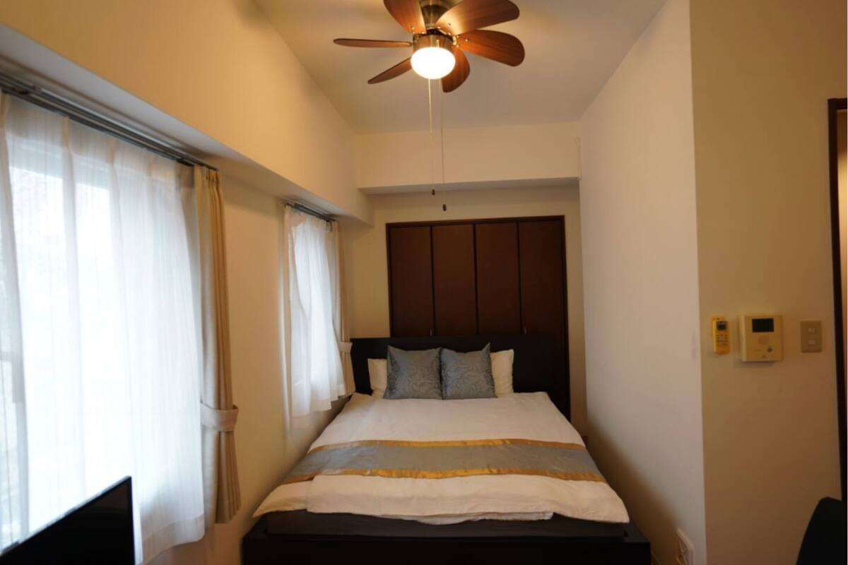 A bedroom with a bed, curtains, a ceiling fan, and a wall-mounted thermostat.