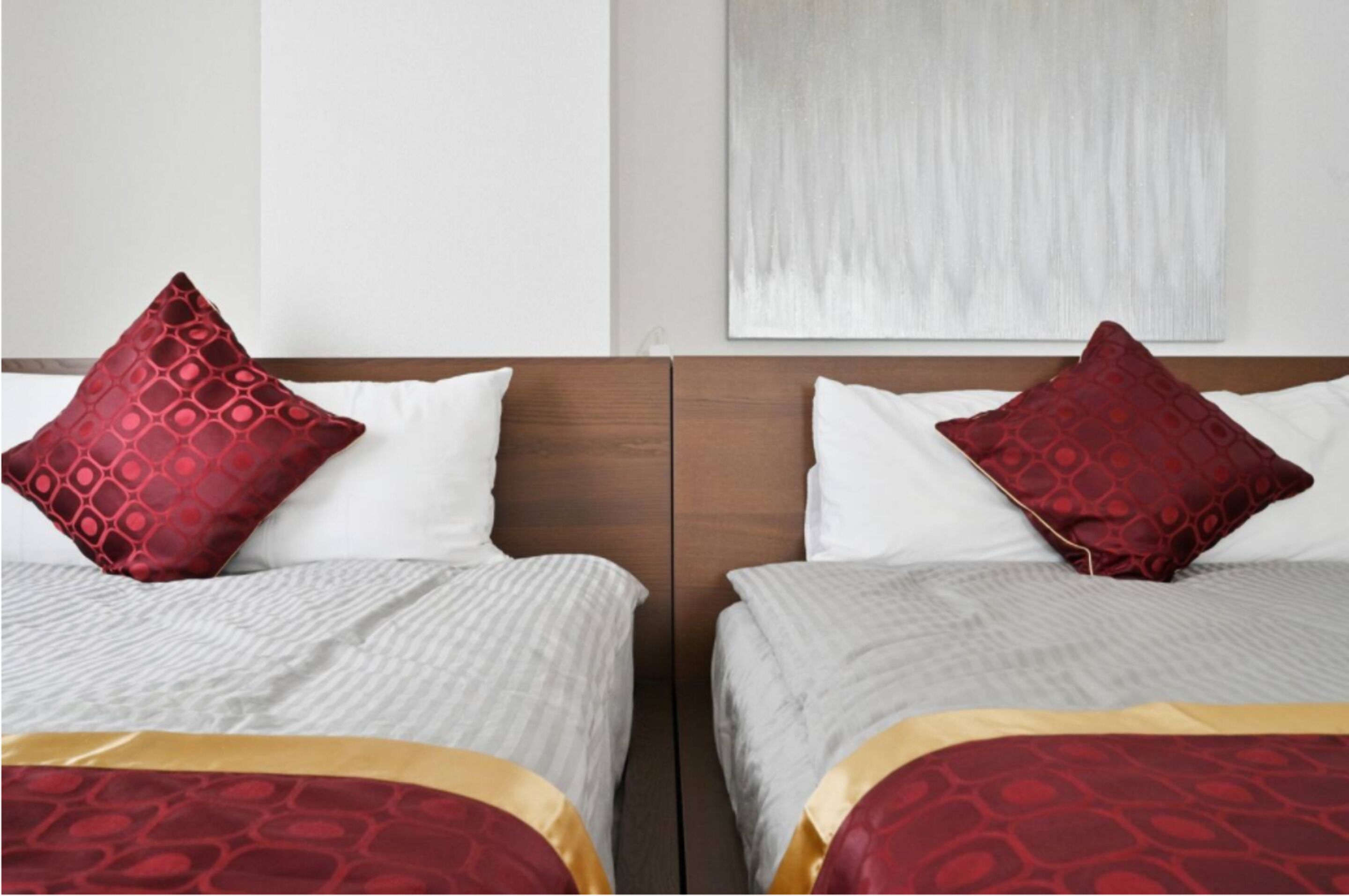 Two beds with white bedding and red patterned pillows, a wooden headboard, and a plain wall with a textured artwork above the beds.