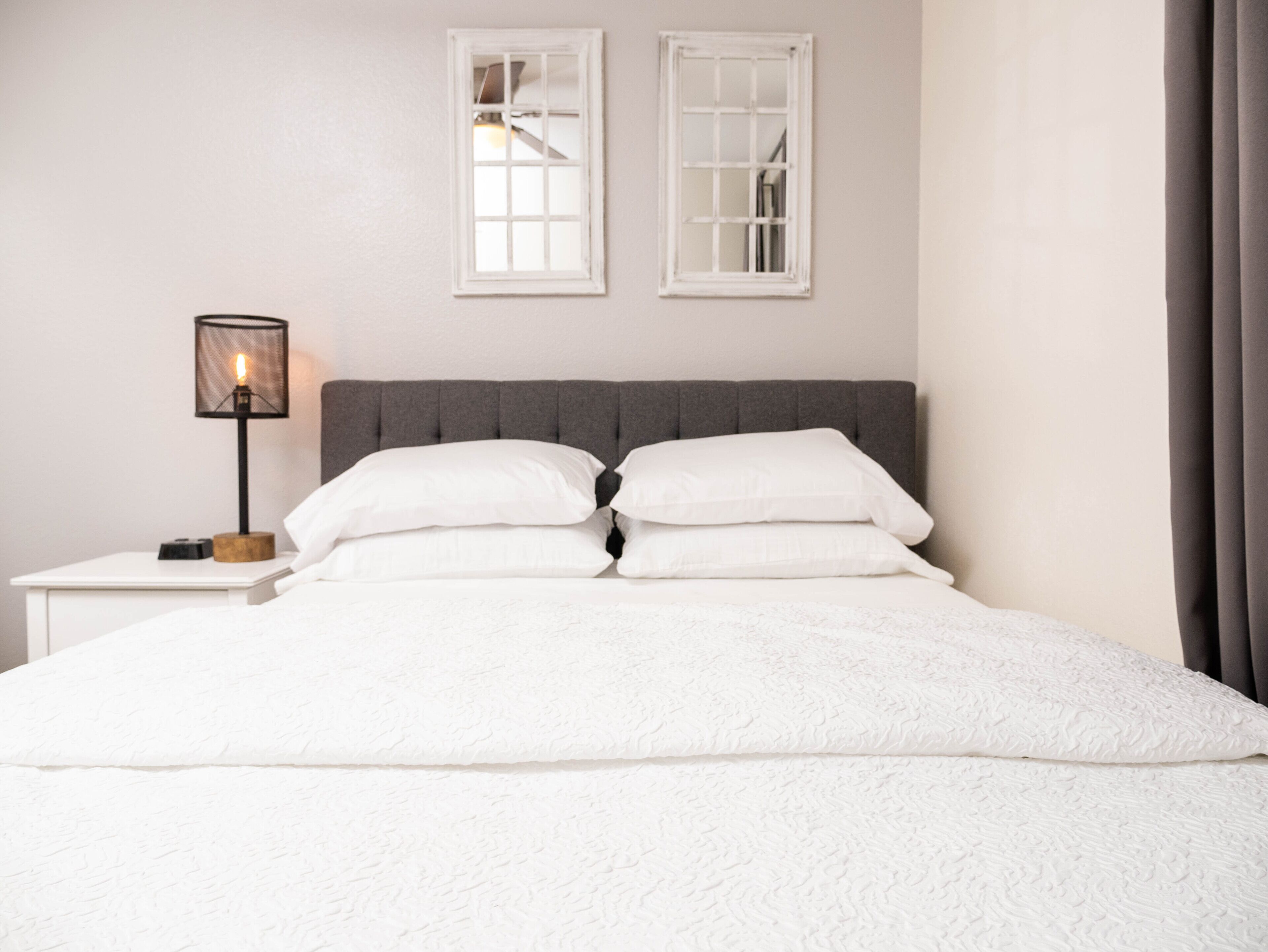 Una cama bien tendida con sábanas blancas y dos almohadas, una lámpara de noche y dos ventanas con marcos blancos.