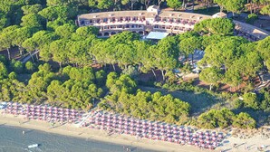 An aerial view of a coastal hotel with a large building surrounded by dense trees, a beach with numerous umbrellas, and a small boat on the water. - Stunning apartment in Puntone with WiFi (Puntone)