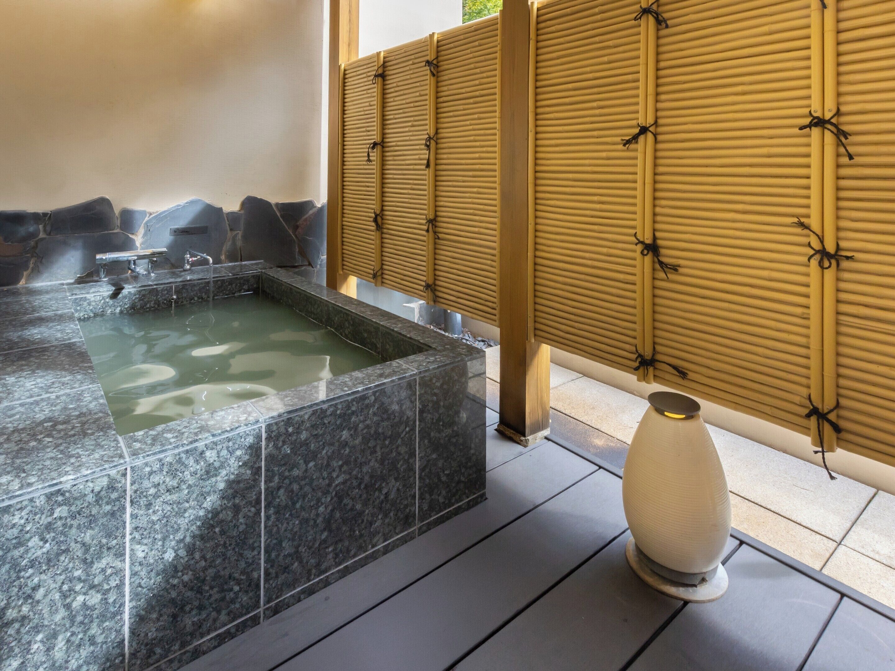 A hot tub with a stone surround, a bamboo screen, and a white vase on a tiled floor.