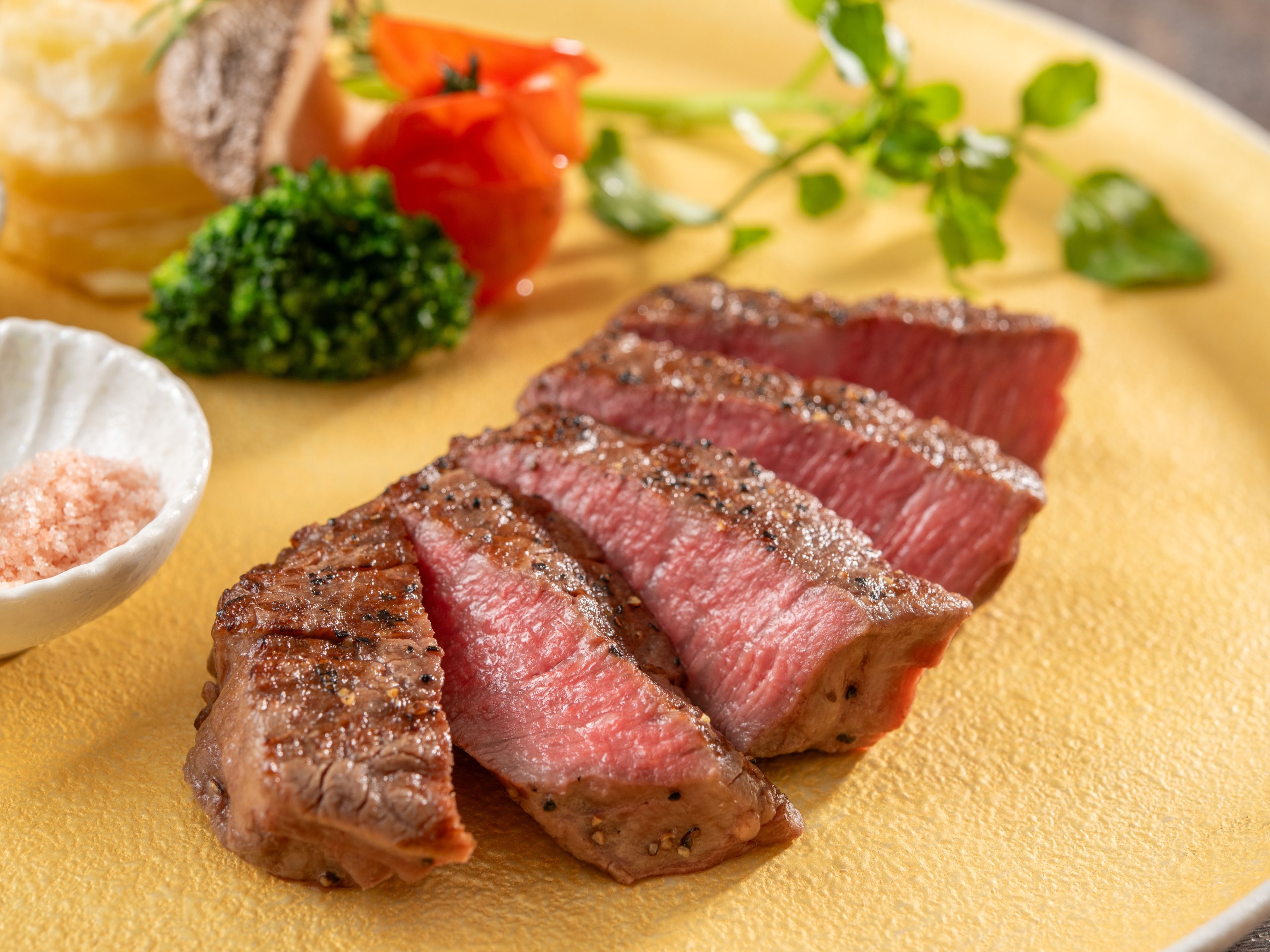 A plate of sliced steak with a side of vegetables and a small bowl of salt.